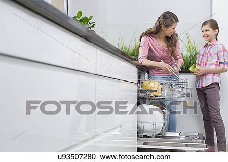 Stock Image - Happy mother and daughter placing glasses in dishwasher at kitchen. Fotosearch
