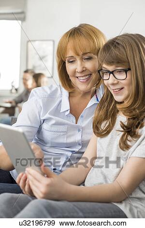 Happy mother and daughter using tablet PC with family sitting in background at home View Large Photo Image Stock Photo - Happy mother and daughter using tablet PC with family sitting in background at home. Fotosearch