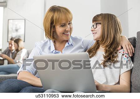 Happy mother and daughter with laptop while family sitting in background at home View Large Photo Image Stock Image - Happy mother and daughter with laptop while family sitting in background at home. Fotosearch