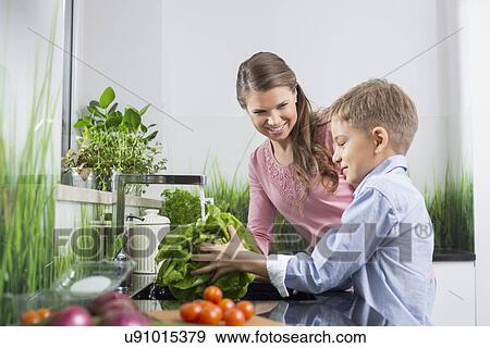 Stock Photo - Happy mother looking at son washing vegetables in kitchen. Fotosearch