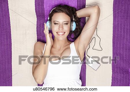 Stock Photograph - Happy woman listening to music through MP3 player using headphones while lying on picnic blanket. Fotosearch