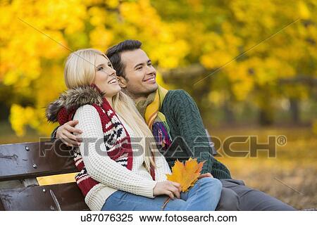 Stock Photography - Happy young couple looking away while sitting on park bench during autumn. Fotosearch