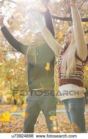 Stock Photo - Happy young couple with arms raised enjoying falling autumn leaves in park. Fotosearch