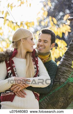 Stock Image - Happy young man hugging woman while leaning on tree trunk during autumn in park. Fotosearch