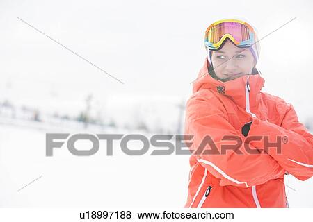 Stock Photo - Happy young woman in warm clothing standing arms crossed on snowy field. Fotosearch