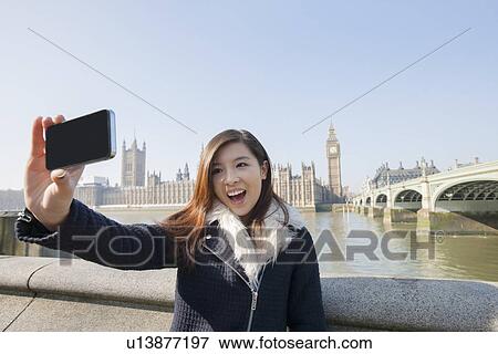 Stock Photo - Happy young woman taking self portrait through cell phone against Big Ben at London, England, UK. Fotosearch