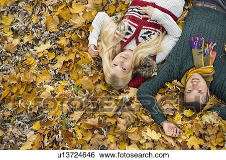 Stock Photograph - High angle portrait of young couple lying on autumn leaves at park. Fotosearch