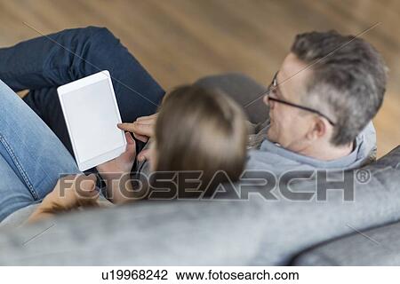 Stock Image - High angle view of father and daughter using digital tablet on sofa. Fotosearch