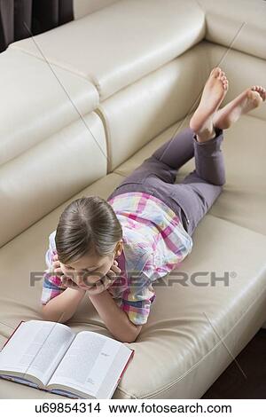 High angle view of girl reading book while lying on sofa at home View Large Photo Image Picture - High angle view of girl reading book while lying on sofa at home. Fotosearch