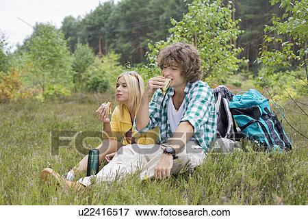 Hiking couple eating sandwiches while relaxing in field View Large Photo Image Stock Photo - Hiking couple eating sandwiches while relaxing in field. Fotosearch