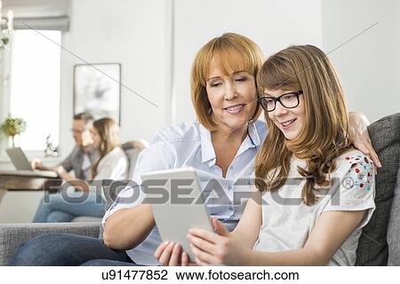 Loving mother and daughter using tablet PC with family sitting in background at home View Large Photo Image Stock Image - Loving mother and daughter using tablet PC with family sitting in background at home. Fotosearch