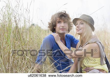 Loving young couple looking away while sitting in field View Large Photo Image Stock Photo - Loving young couple looking away while sitting in field. Fotosearch