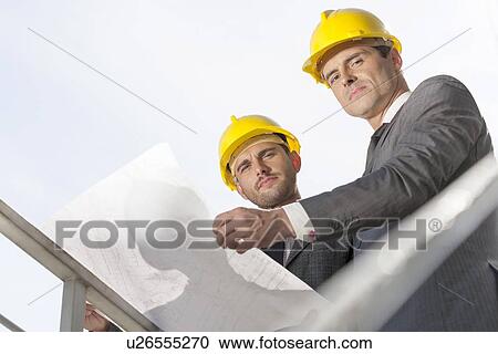 Stock Image - Low angle portrait of confident young businessmen holding blueprint against clear sky. Fotosearch