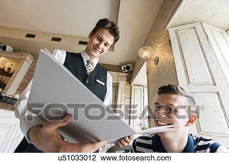 Stock Image - Low angle view of waiter showing menu to male customer in restaurant. Fotosearch