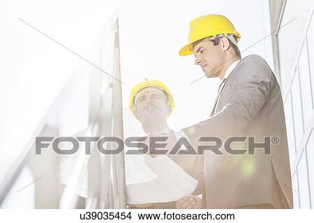 Low angle view of young businessman in hard hats reviewing blueprint on stairway View Large Photo Image Picture - Low angle view of young businessman in hard hats reviewing blueprint on stairway. Fotosearch