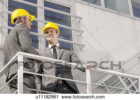 Stock Image - Low angle view of young male architect with coworker discussing on stairway. Fotosearch