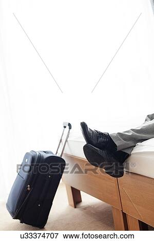 Stock Photo - Low section of businessman lying on bed beside luggage in hotel room. Fotosearch