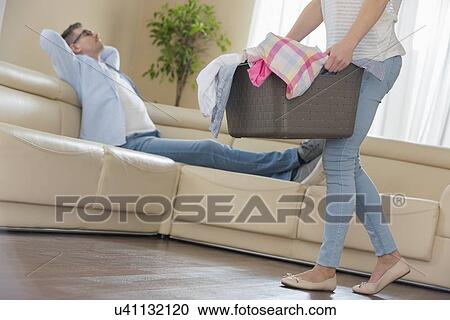 Stock Image - Low section of woman walking with laundry basket while man relaxing on sofa in background. Fotosearch