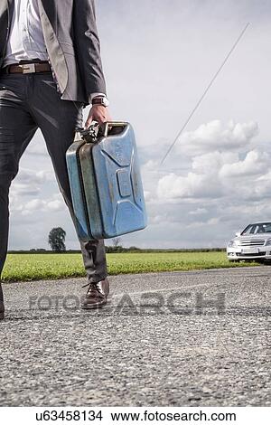 Picture - Low section of young businessman carrying gasoline can with broken car in background at countryside. Fotosearch