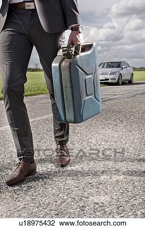 Stock Image - Low section of young businessman carrying gas can with broken car in background at countryside. Fotosearch