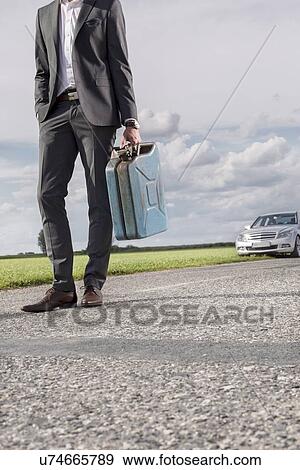 Stock Photo - Low section of young businessman carrying petrol can with broken car in background at countryside. Fotosearch