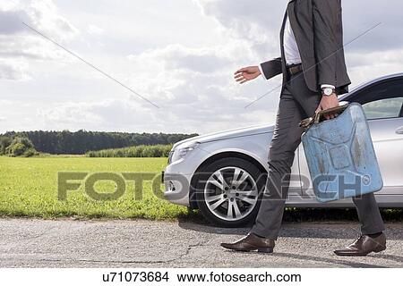 Picture - Low section of young businessman with gas can walking by broken down car at countryside. Fotosearch