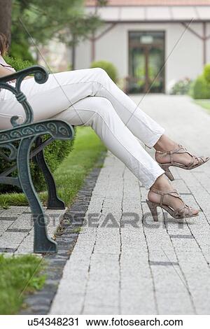 Stock Image - Low section of young woman with high heels sitting on park bench. Fotosearch