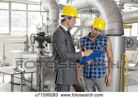 Stock Image - Male architect with worker discussing over clipboard in industry. Fotosearch