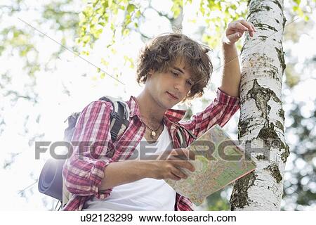 Stock Photo - Male backpacker reading map while leaning on tree trunk in forest. Fotosearch