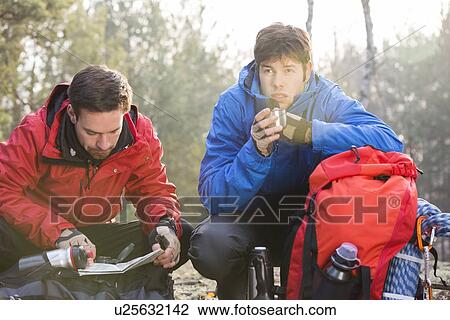 Stock Image - Male hiker reading map while friend having coffee in forest. Fotosearch