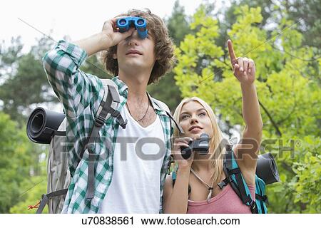 Stock Image - Male hiker using binoculars while girlfriend showing something in forest. Fotosearch