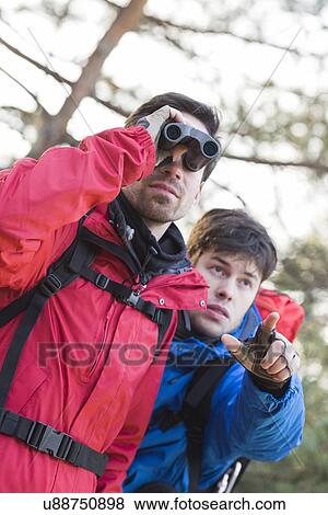 Stock Photo - Male hiker using binoculars while friend showing him something in forest. Fotosearch