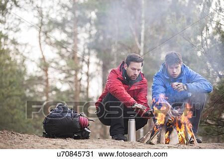 Stock Image - Male hikers warming hands at campfire in forest. Fotosearch