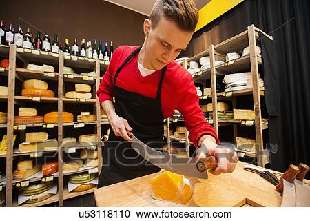 Stock Image - Male salesperson cutting cheese in store. Fotosearch