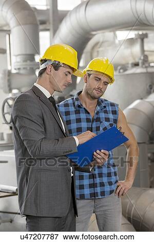 Stock Photo - Manual worker with supervisor discussing over clipboard in industry. Fotosearch