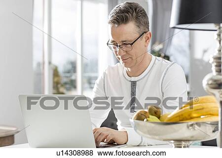 Stock Photo - Mature man using laptop at table in house. Fotosearch