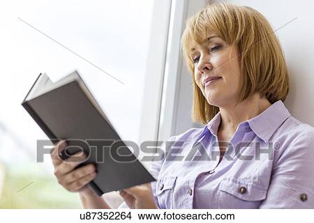 Mature woman reading book by window at home View Large Photo Image Picture - Mature woman reading book by window at home. Fotosearch