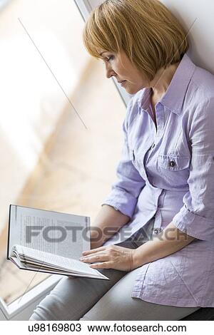 Mature woman reading book while sitting by window at home View Large Photo Image Stock Image - Mature woman reading book while sitting by window at home. Fotosearch