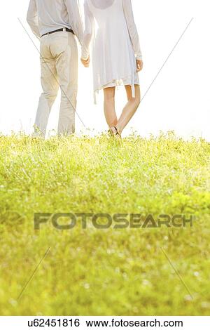 Midsection of couple holding hands while standing on grass against sky View Large Photo Image Stock Photograph - Midsection of couple holding hands while standing on grass against sky. Fotosearch