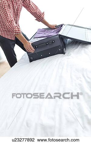 Stock Image - Midsection of young businessman packing suitcase in bedroom. Fotosearch