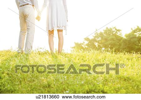 Midsection rear view of couple holding hands while standing on grass against sky View Large Photo Image Stock Image - Midsection rear view of couple holding hands while standing on grass against sky. Fotosearch