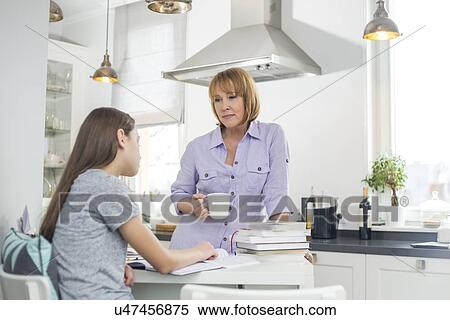 Stock Photography - Mother having coffee while looking at daughter studying in kitchen. Fotosearch