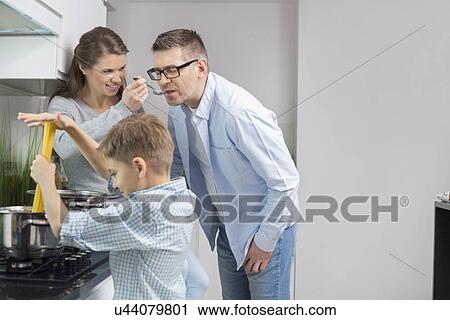 Stock Image - Parents tasting food while son preparing food in kitchen. Fotosearch