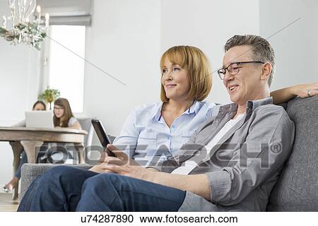 Stock Image - Parents watching TV on sofa with children using laptop in background. Fotosearch