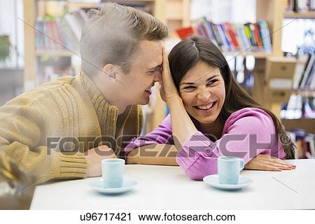 Stock Image - Playful young couple with coffee cups on desk in library. Fotosearch