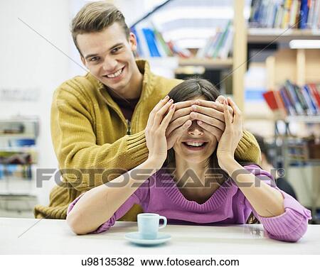 Playful young man covering woman's eyes in library View Large Photo Image Stock Image - Playful young man covering woman's eyes in library. Fotosearch
