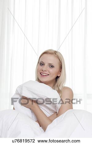 Stock Image - Portrait of attractive young woman covering herself with bedsheet in bed. Fotosearch