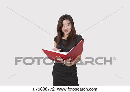 Stock Image - Portrait of beautiful young businesswoman writing notes in folder over white background. Fotosearch
