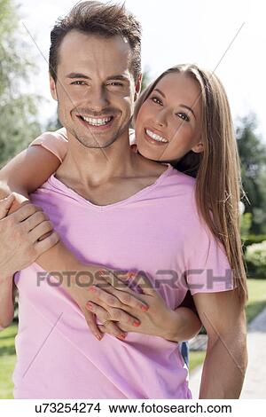 Portrait of beautiful young woman embracing man from behind in park View Large Photo Image Picture - Portrait of beautiful young woman embracing man from behind in park. Fotosearch