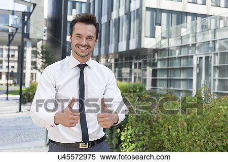 Portrait of confident businessman showing thumbs up outside office building View Large Photo Image Stock Photography - Portrait of confident businessman showing thumbs up outside office building. Fotosearch